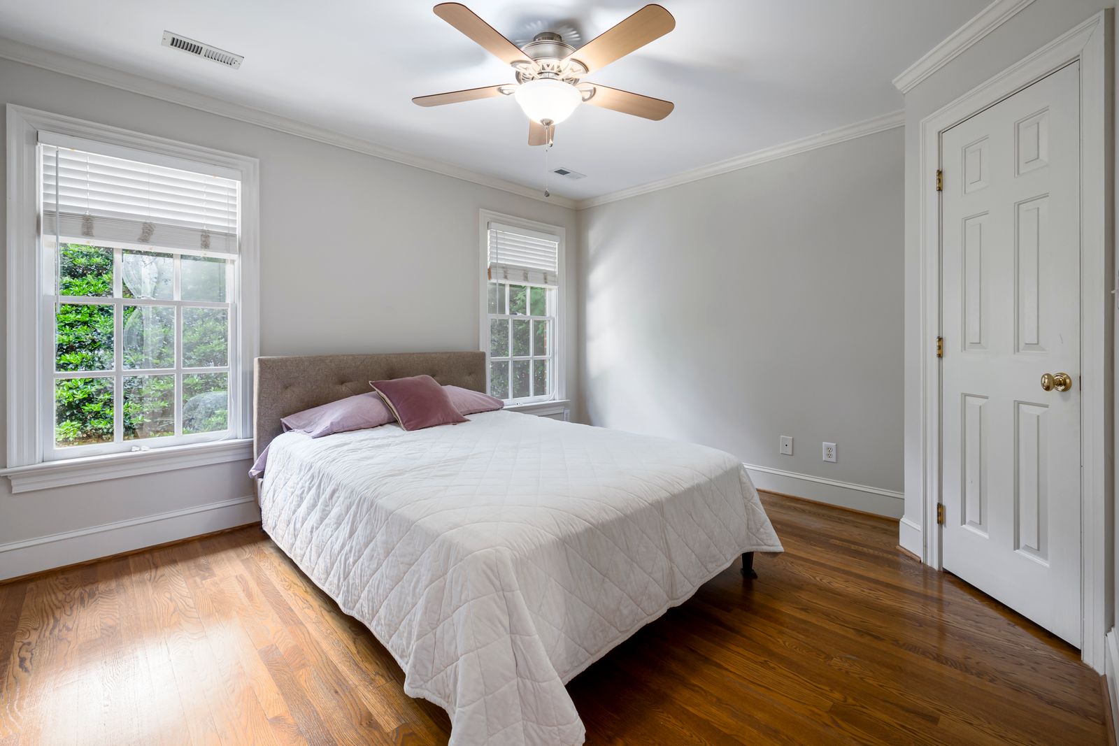 A bedroom with a quilted white bedspread, a ceiling fan, tall windows with blinds, and a panelled white door