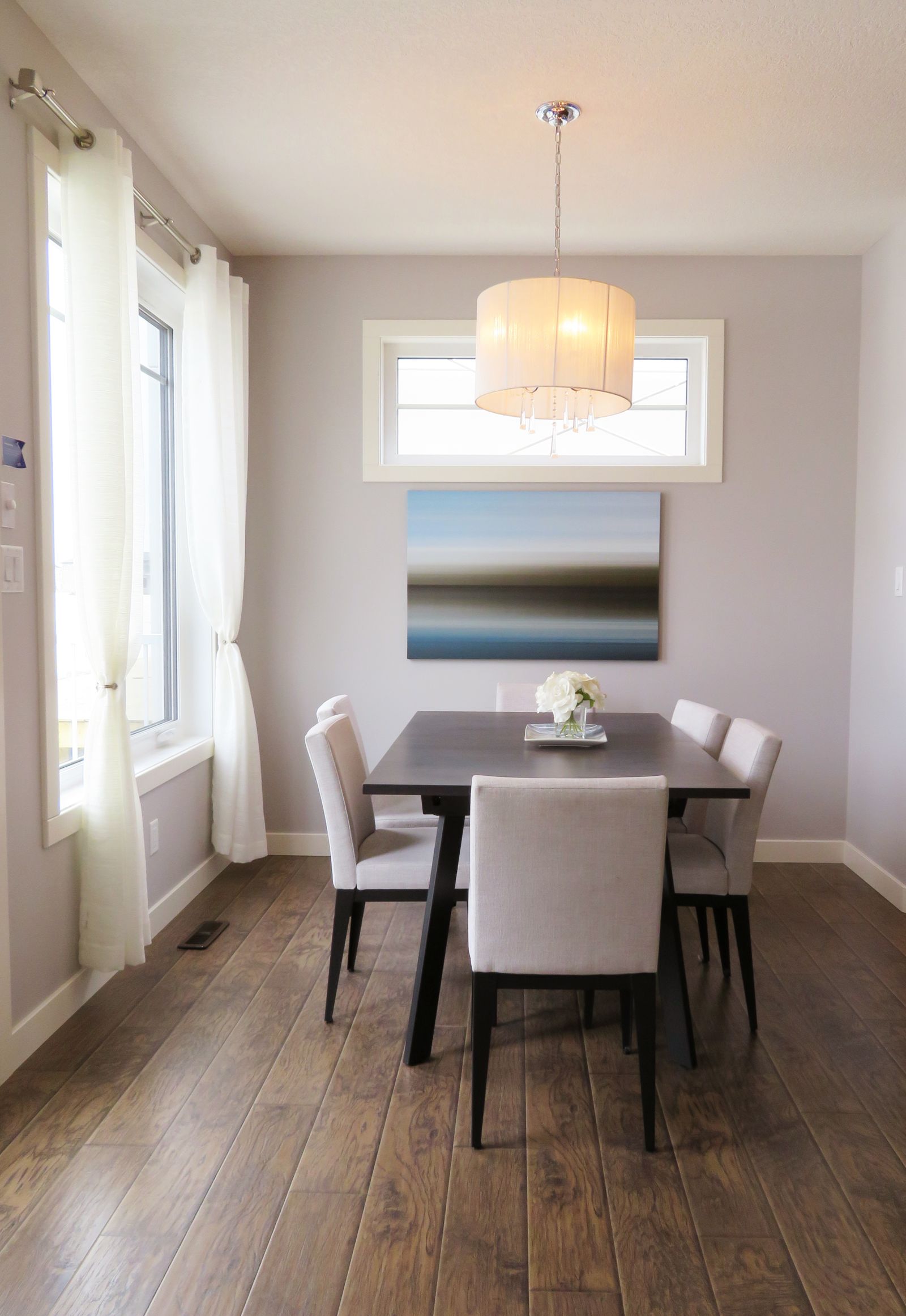 A dining room with a dark wood table, upholstered chairs, a drum pendant light, and a framed horizon painting on a lavender-grey wall