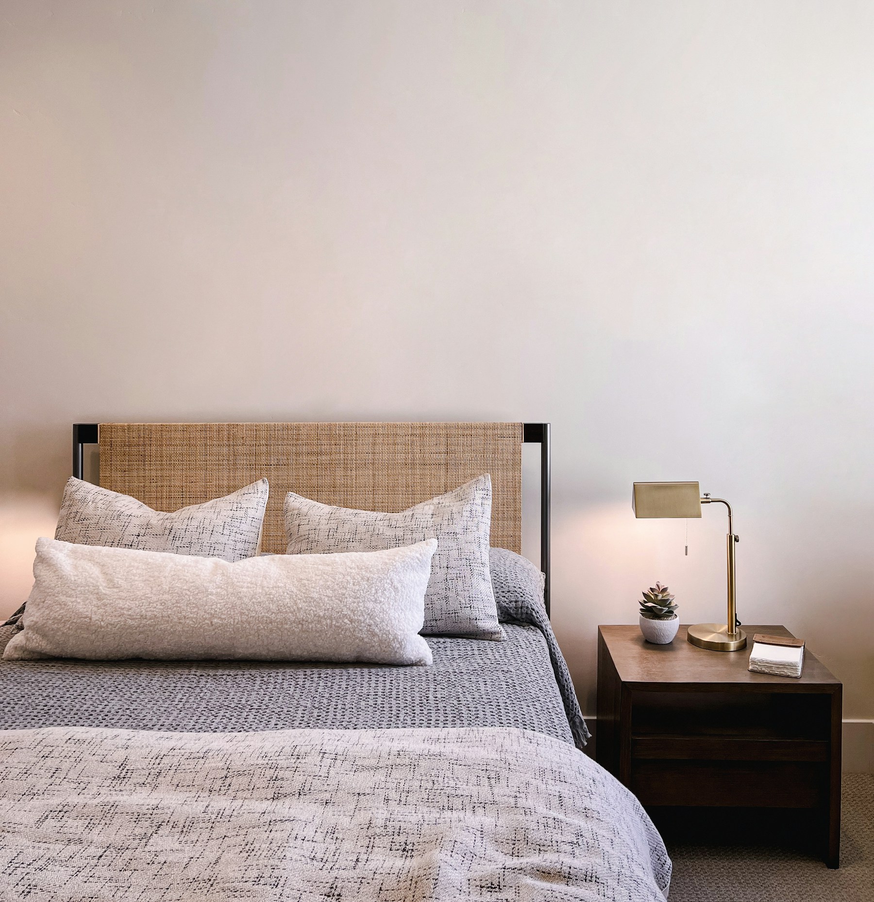 A close-up of a woven rattan bed headboard with grey textured pillows and a knit throw, beside a walnut bedside table holding a small potted succulent and a brass desk lamp