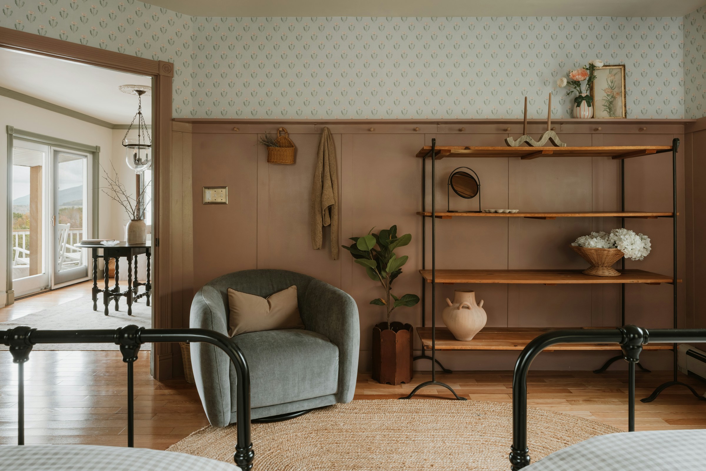 A bedroom with a black iron bed frame, floral wallpaper above wood wainscoting, a curved sage bouclé armchair, a black metal étagère styled with pottery, and a jute rug