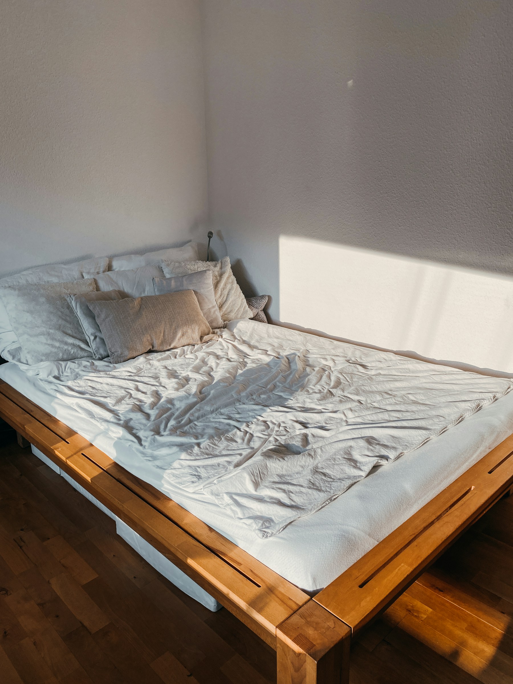 A simple wood-slat bed frame with rumpled white bedding, lit by a sharp diagonal shaft of afternoon sun across the corner wall