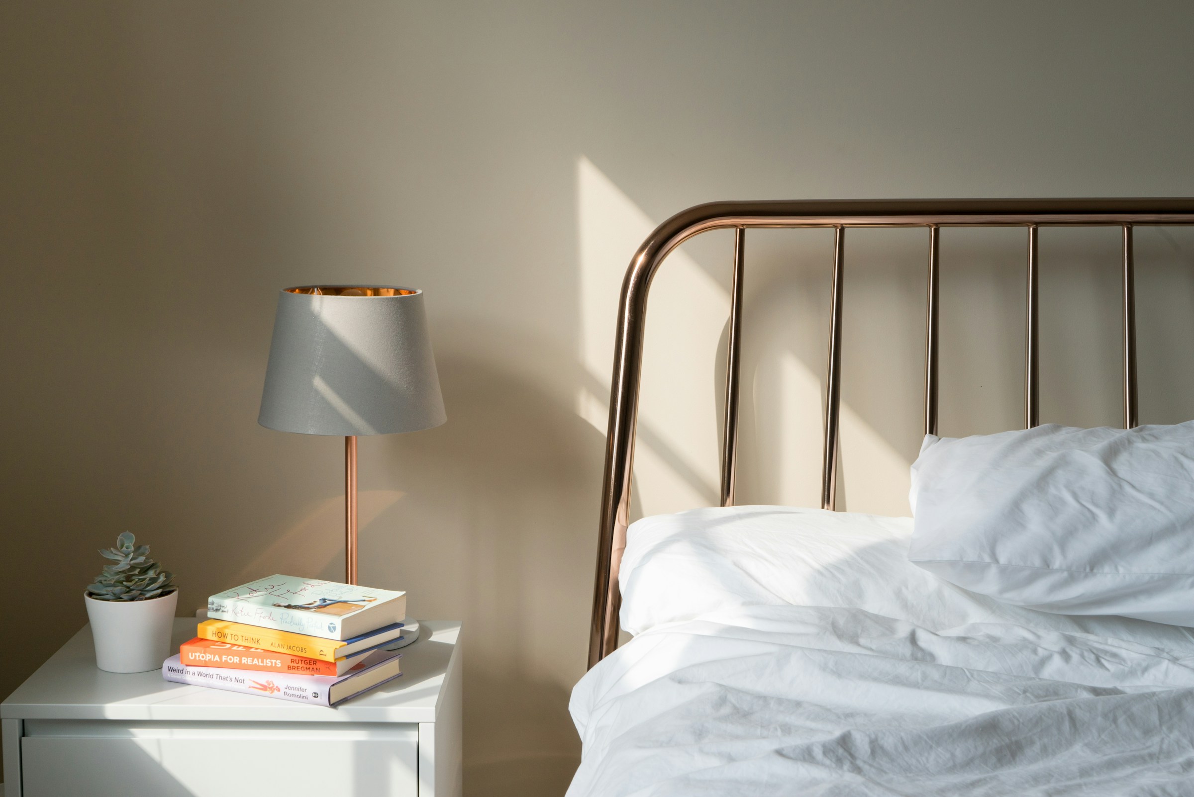 A bed with crumpled white linens raked by warm afternoon sunlight, beside a bedside table holding a small succulent, a stack of books, and a copper table lamp with a grey shade