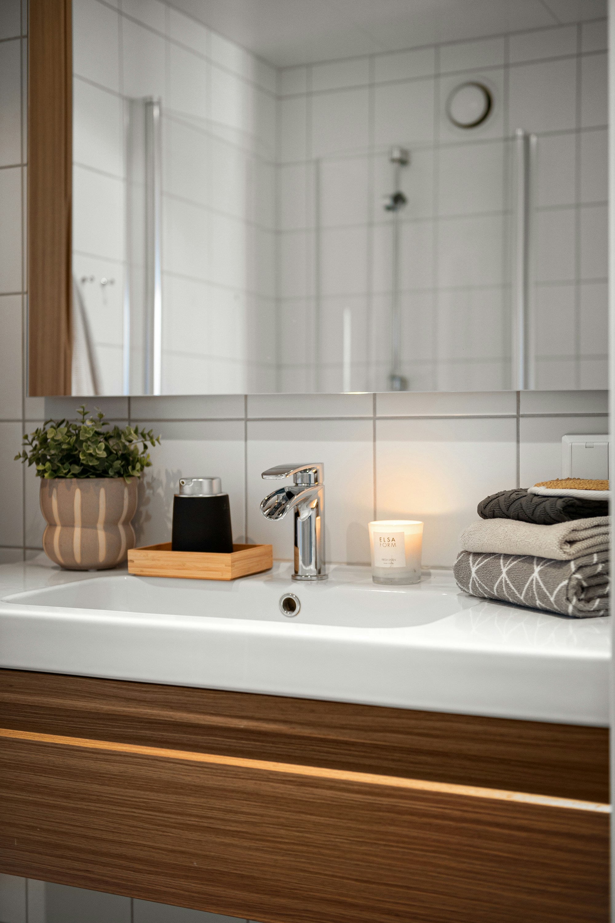An en-suite bathroom vanity with a white ceramic basin set into a walnut wood-grain cabinet, a chrome tap, a lit candle, and folded grey towels, reflected in a mirror showing a glass shower stall