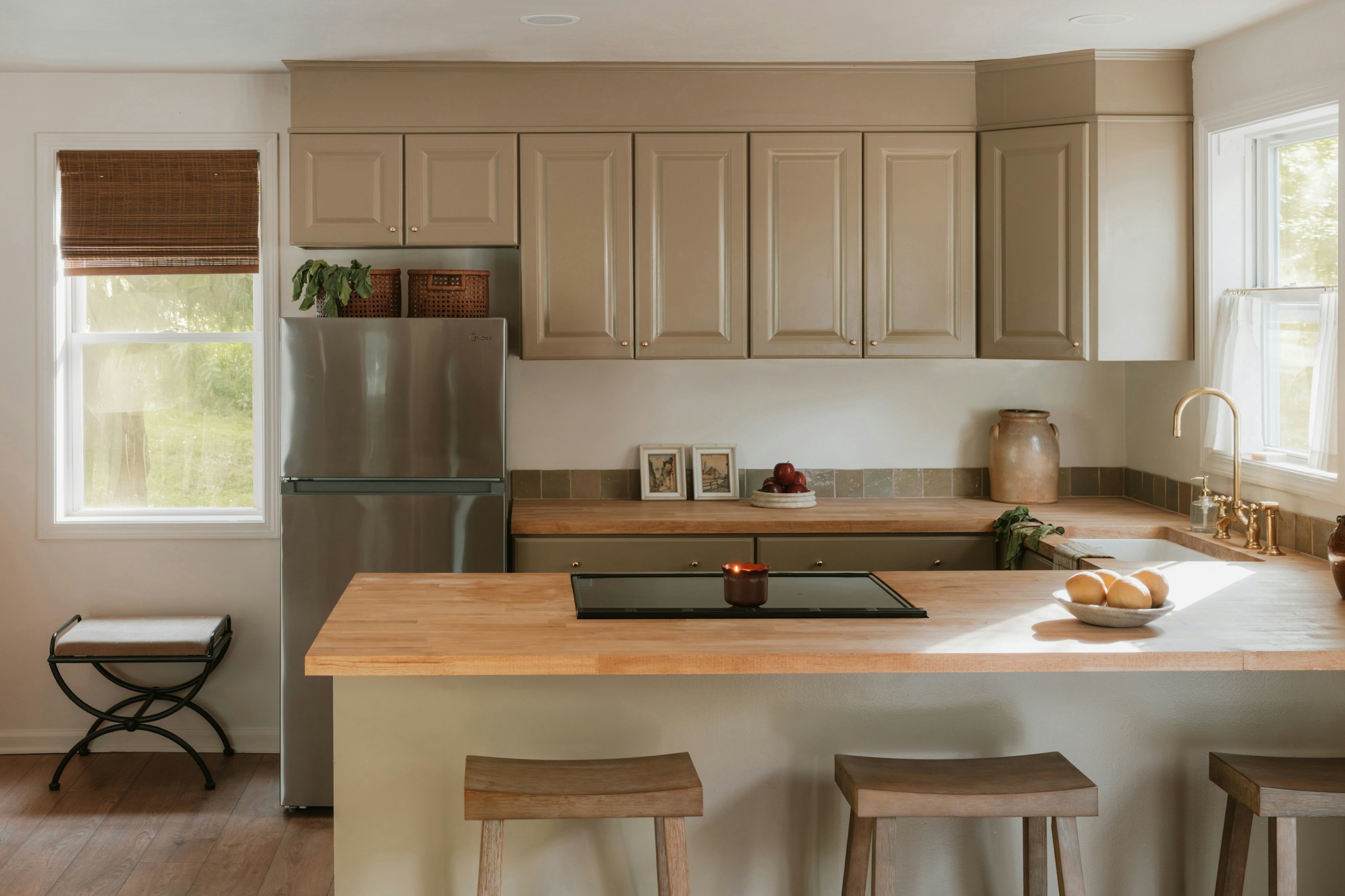 A kitchen with warm olive-toned cabinetry, a natural oak butcher-block island, a brass gooseneck tap, a stoneware jug and bowl of lemons, and three wood counter stools lit by a bright side window