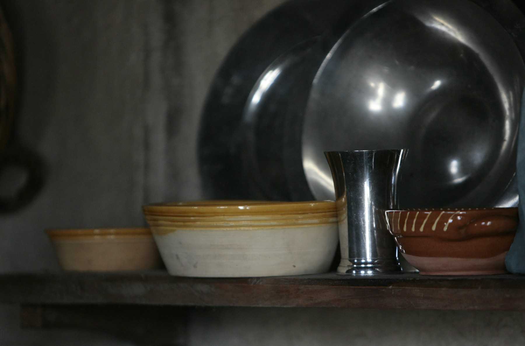 A quiet still life on a wooden shelf: pewter chargers leaning upright behind a mustard-rimmed cream ceramic bowl and a rust-glazed pottery bowl