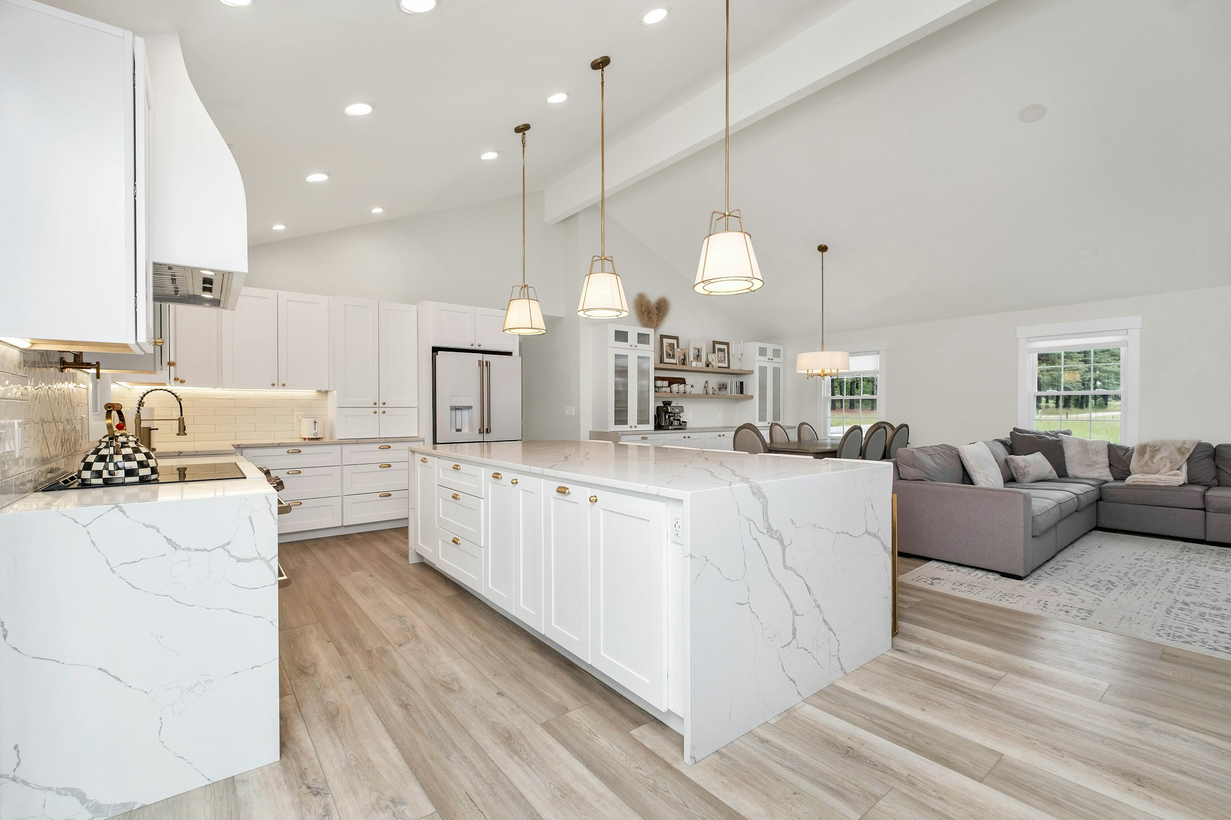A large open-plan kitchen with a white marble waterfall-edge island, brass pendant lights, white shaker cabinetry, and a sofa visible in the living area beyond, floored in light wood