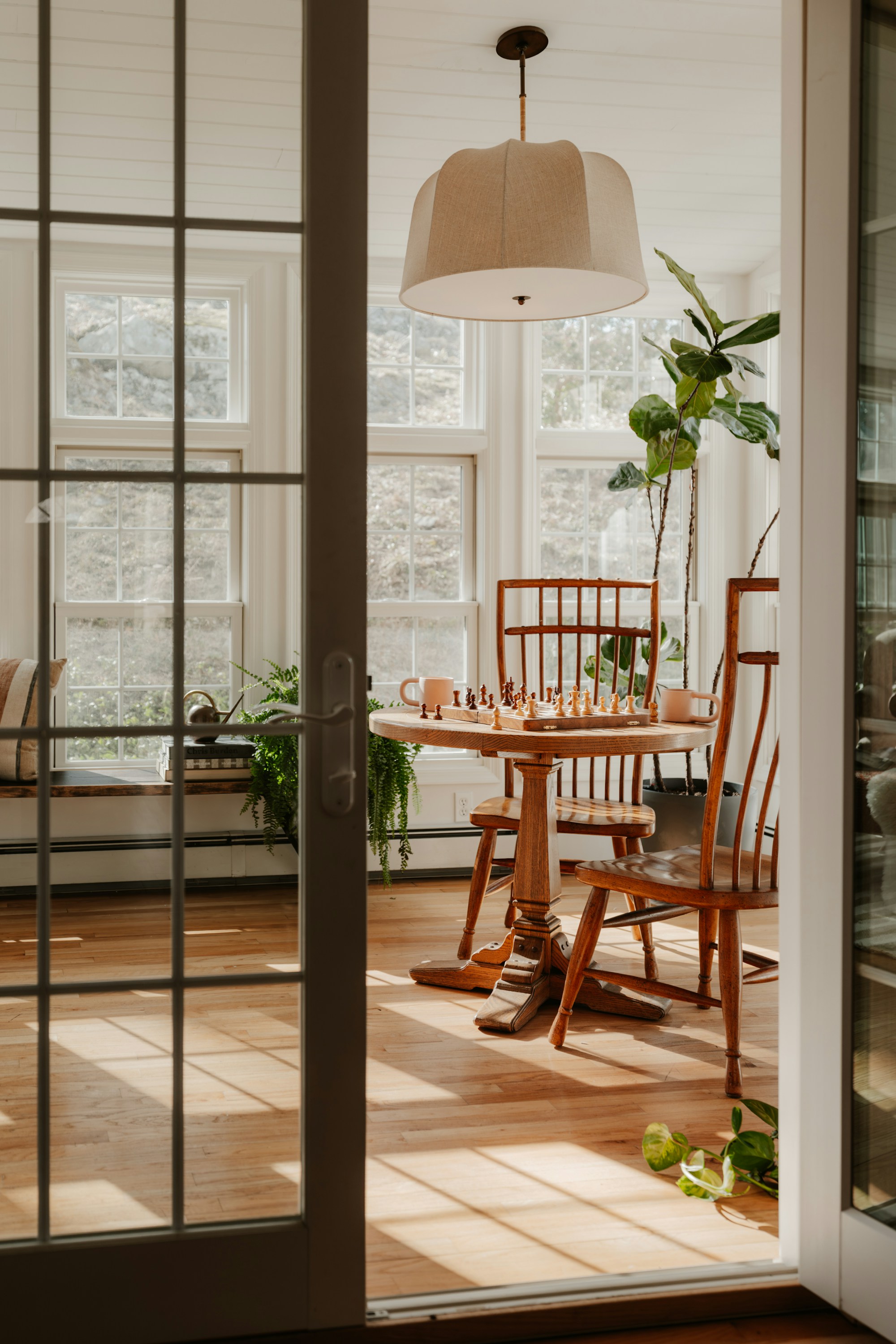 Viewed through a glass-paned French door, a sunlit breakfast nook with a round oak pedestal table, wooden spindle chairs, a linen drum pendant shade, and potted plants against tall multi-pane windows