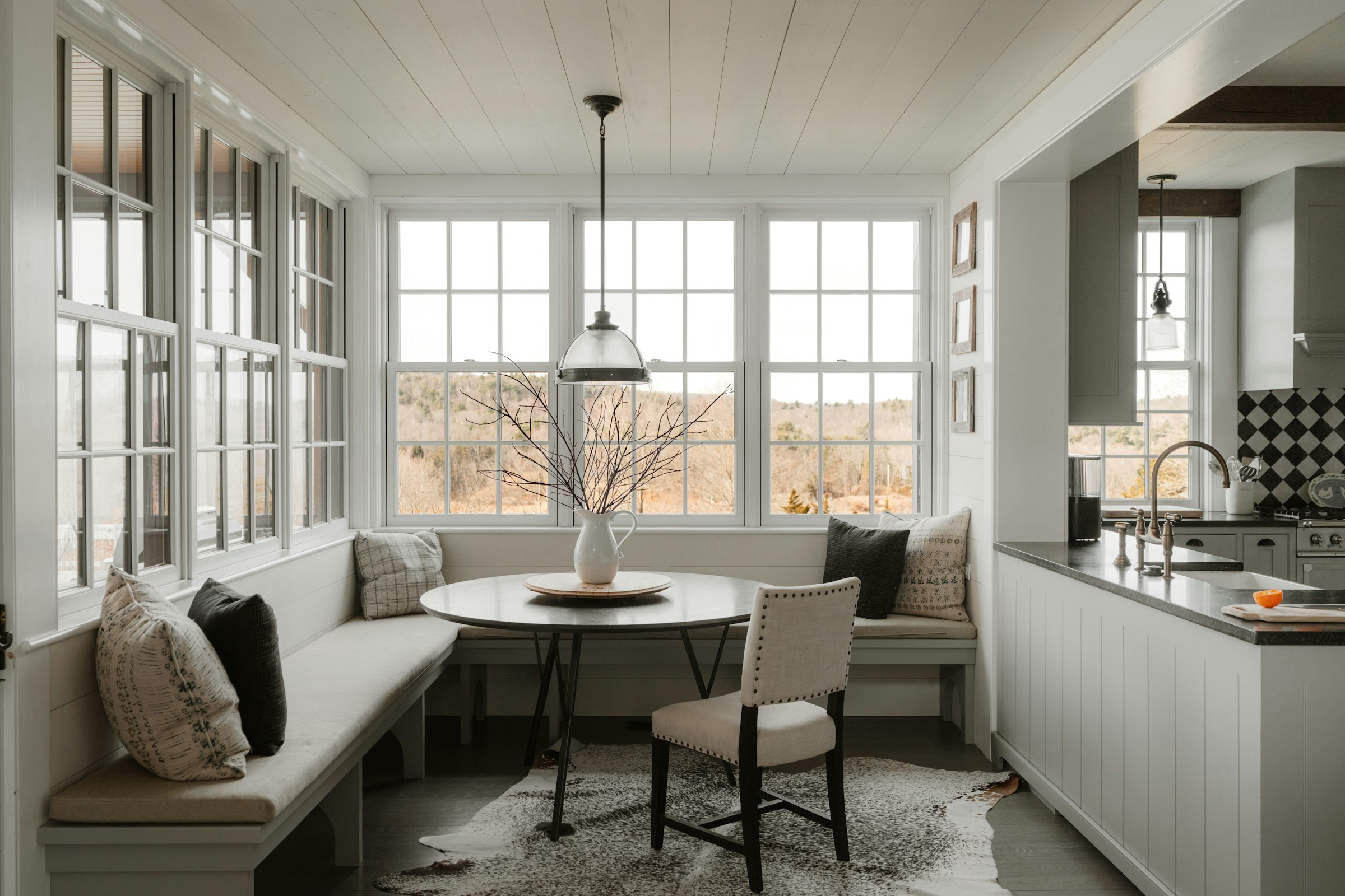 A white shiplap-walled breakfast nook with an L-shaped built-in bench, a round pedestal table with a ceramic pitcher, a dome pendant light, and a cowhide rug opening into the kitchen beyond