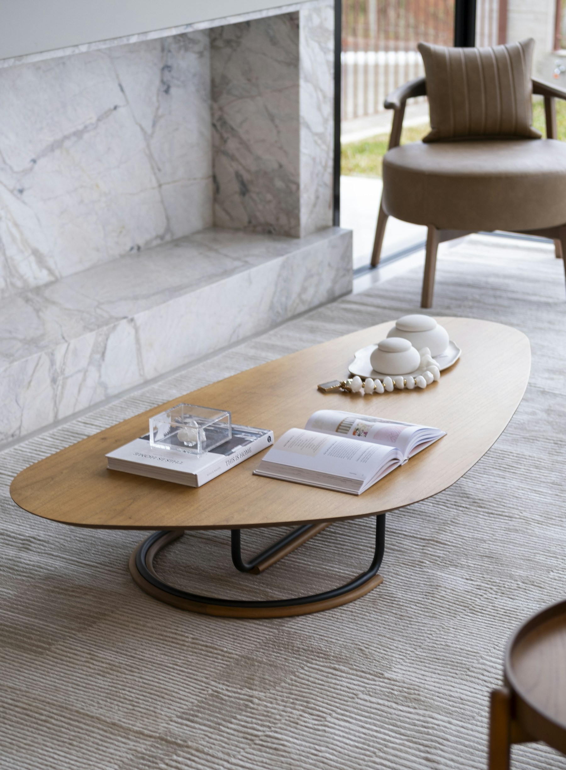 A sculptural oval wood coffee table with a looped metal base, styled with an open book, stacked design books, and small white ceramic vessels beside a marble fireplace surround
